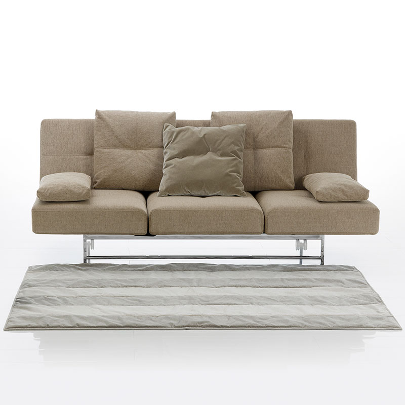 Hellbraunes Sofa mit dreigeteilter Sitzfläche. Statt Armlehnen liegen auf dem Sofa flache, rechteckige Kissen. An der Rückenlehne lehnen drei große, rechteckige Kissen.