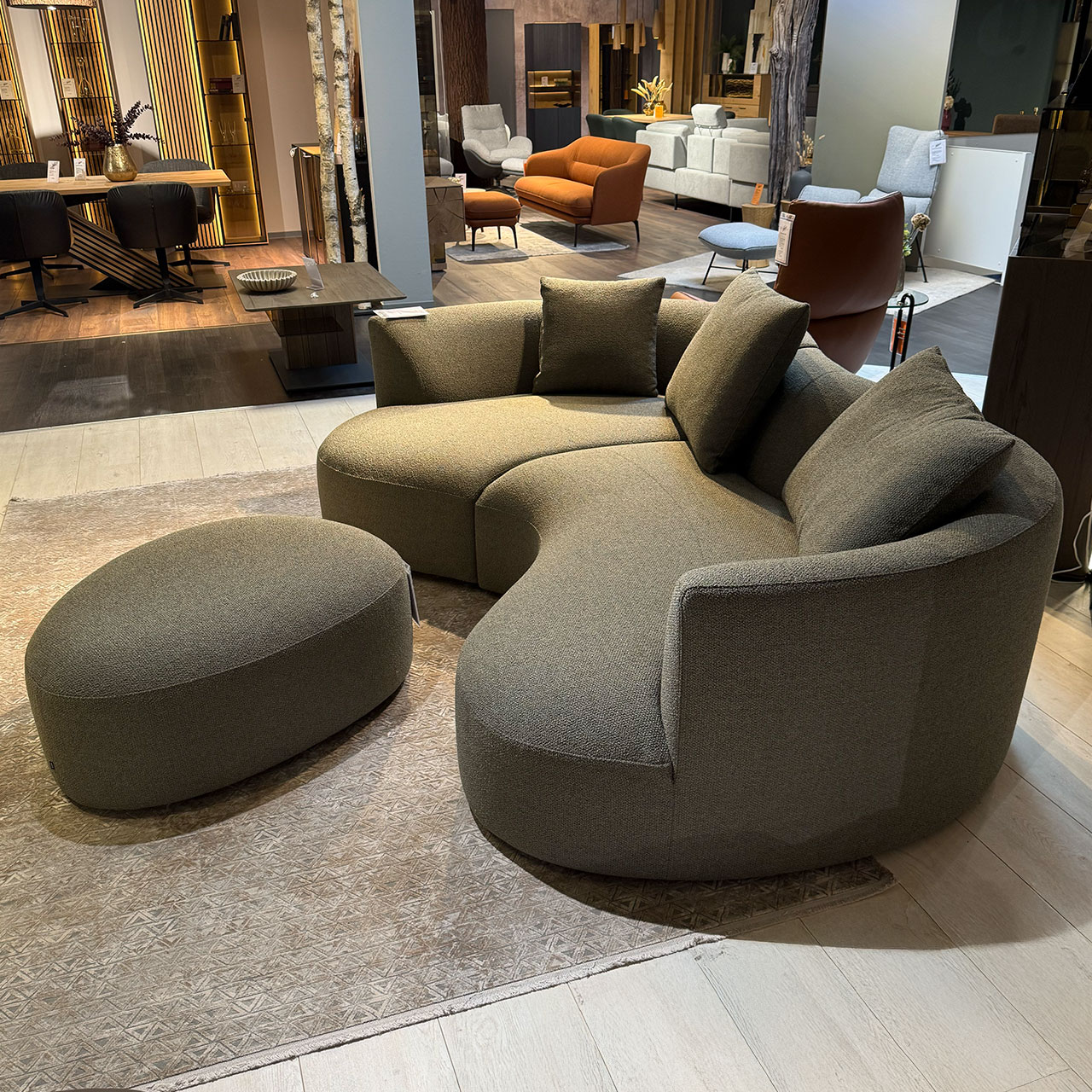 Organisch geformtes Sofa aus graugrünem Stoff mit Liege- und Sitzteil mit einem kleinen und zwei großen Kissen, davor ein ovaler Hocker