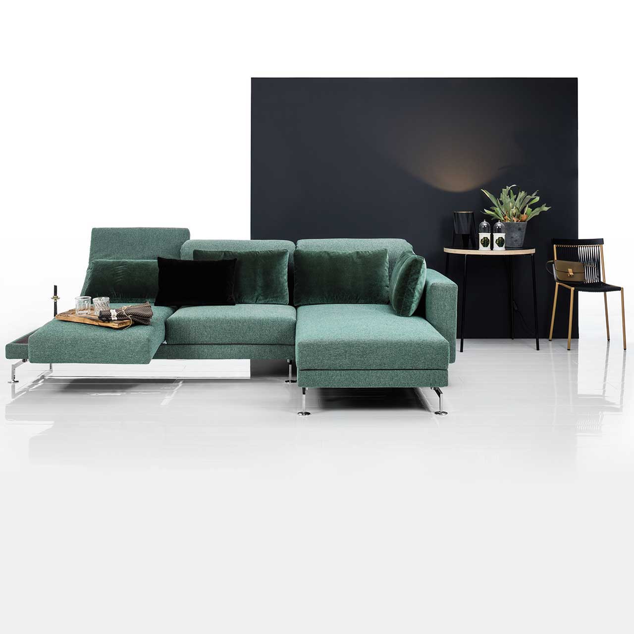 Eckgarnitur mit Longchair rechts in grünem Stoff. Rechts neben der Armlehne befindet sich eine Ablagefläche in dunklem Holz. Die Armlehne ist zur Relaxposition vorgescchwenkt, die Rückenlehne ist abgesenkt.