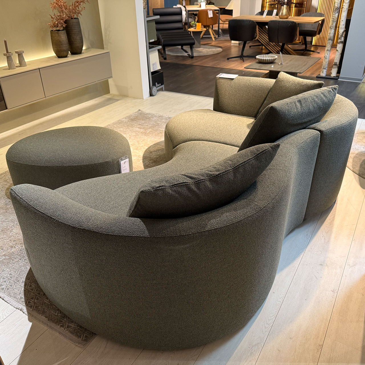 Organisch geformtes Sofa aus graugrünem Stoff mit Liege- und Sitzteil mit einem kleinen und zwei großen Kissen, davor ein ovaler Hocker