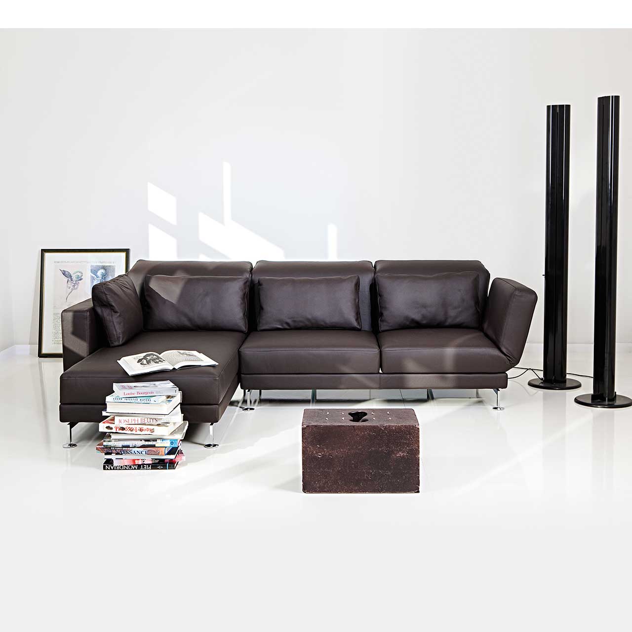 Eckgarnitur in schwarzem Leder mit Longchair links. Vor dem Longchair steht ein Stapel Bücher, links hinter der Garnitur steht ein gerahmtes Bild an die Wand gelehnt. Vor der Garnitur steht ein quadratisches Kunstelement..