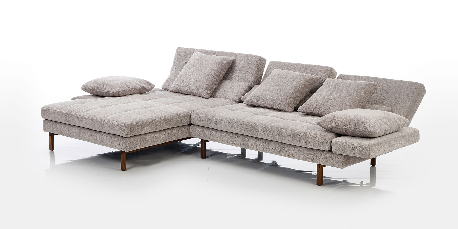 Eine Eckgarnitur in sandfarbenem Stoff mit einem Longchair mit abgeklappter Armlehne links sowie einem Anstellsofa mit leicht abgesenkten Rückenlehnen. Auf der Garnitur liegen vier Kissen.