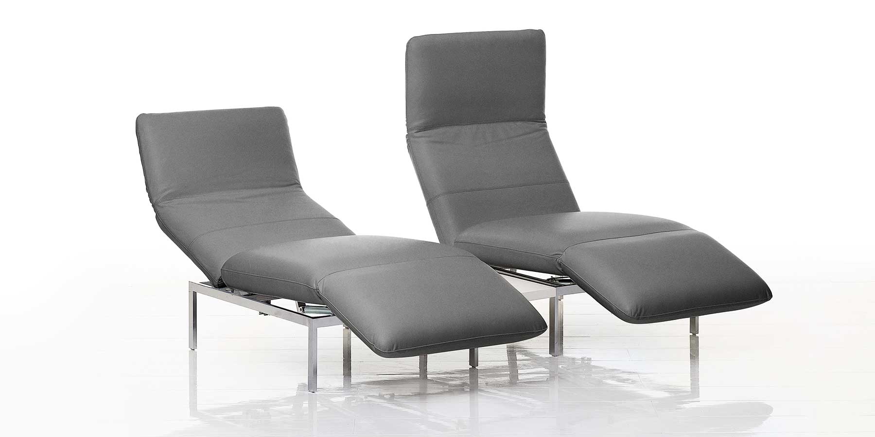 Zwei nebeneinander stehende Multifunktionssessel in grauem Leder. Beide sind zur Relaxposition aufgeschwenkt.