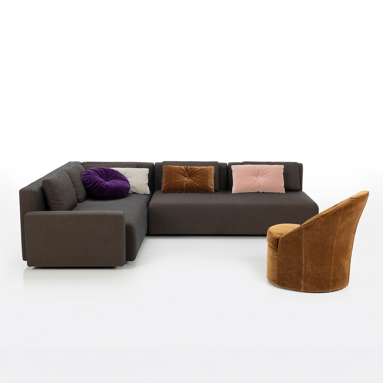 Bodennahes braunes Eckofa mit Longchair links und Drehsofa rechts. Auf dem Sofa liegen drei eckige und ein rundes Kissen. Im Vordergrund steht ein karamellfarbener Sessel.