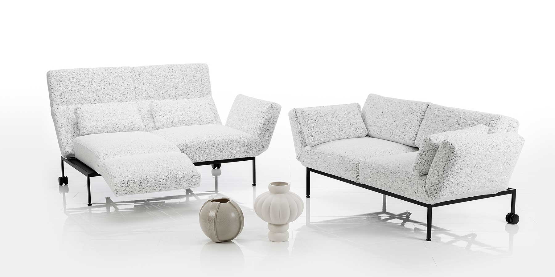 Zwei Sofas mit weißen Stoffbezügen und schwarzem Untergestell. Die linke Armlehne des links stehenden Sofas ist zur Fußstütze vorgeschwenkt.