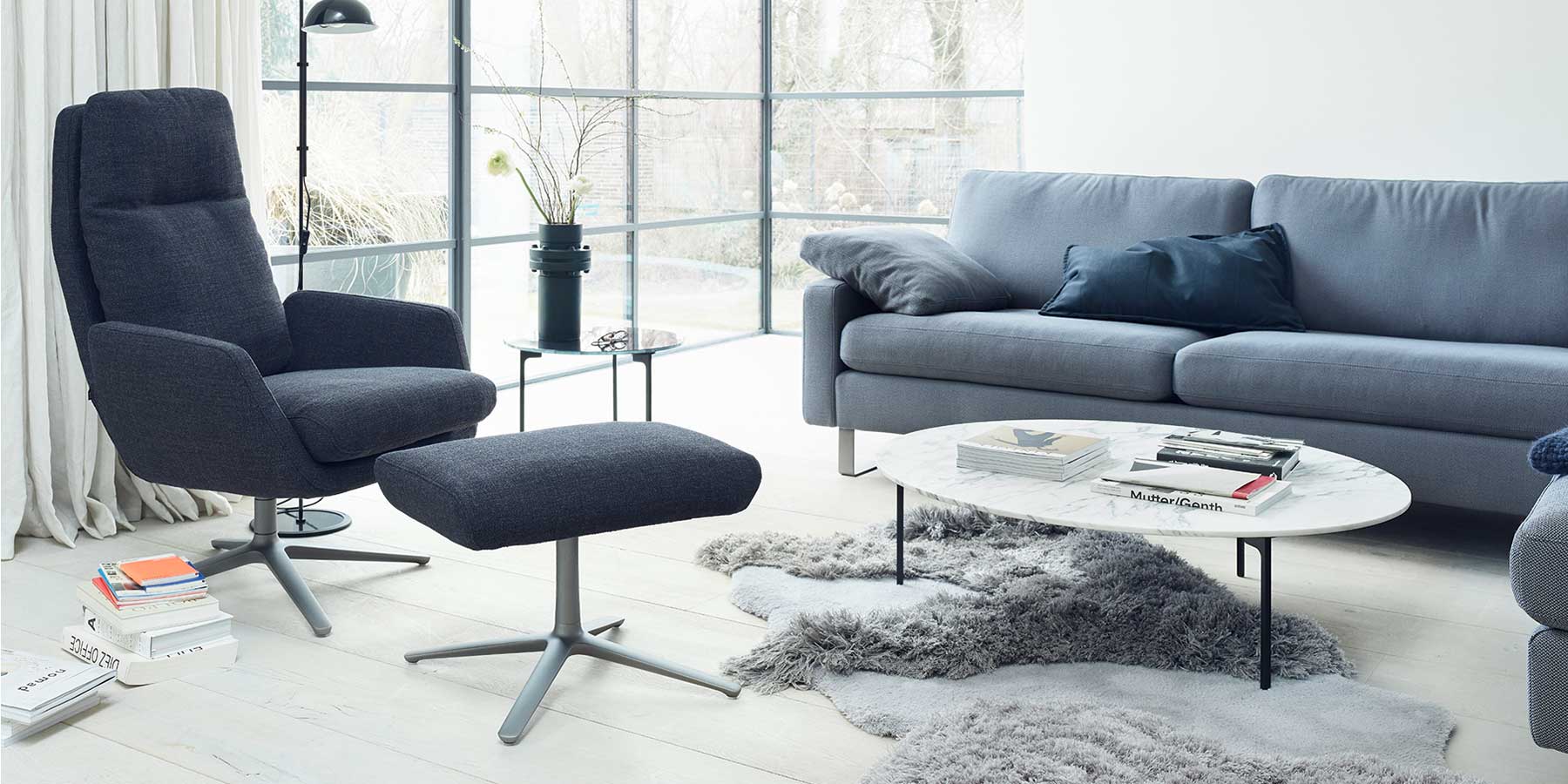 Dunkelblauer COR Cordia Sessel mit Hocker neben einer hellblauen Couch in einem Wohnzimmer