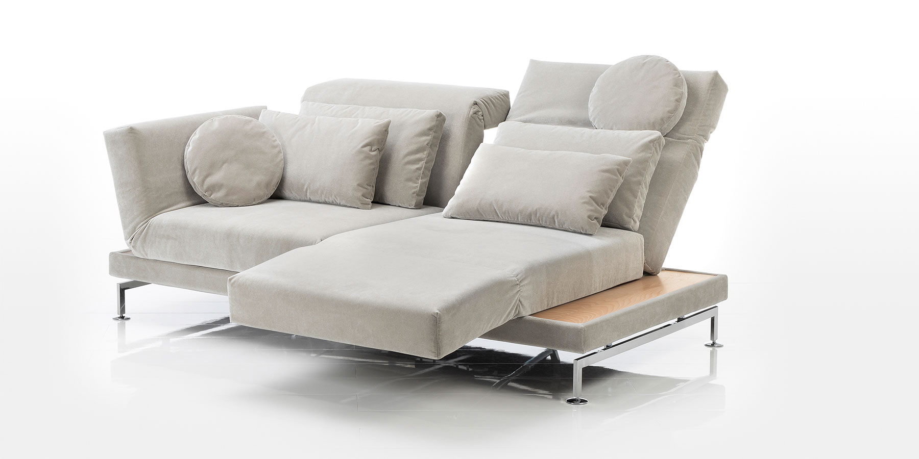 Ein Sofa mit zwei Sitzplätzen in einem dunklen beige. Der rechte Sitz wurde um 90 Grad gedreht und die Armlehne so abgesenkt, dass sie als Fußstütze verwendet werden kann. Auf dem Sofa wurden vier rechteckige sowie zwei runde Kissen drapiert.