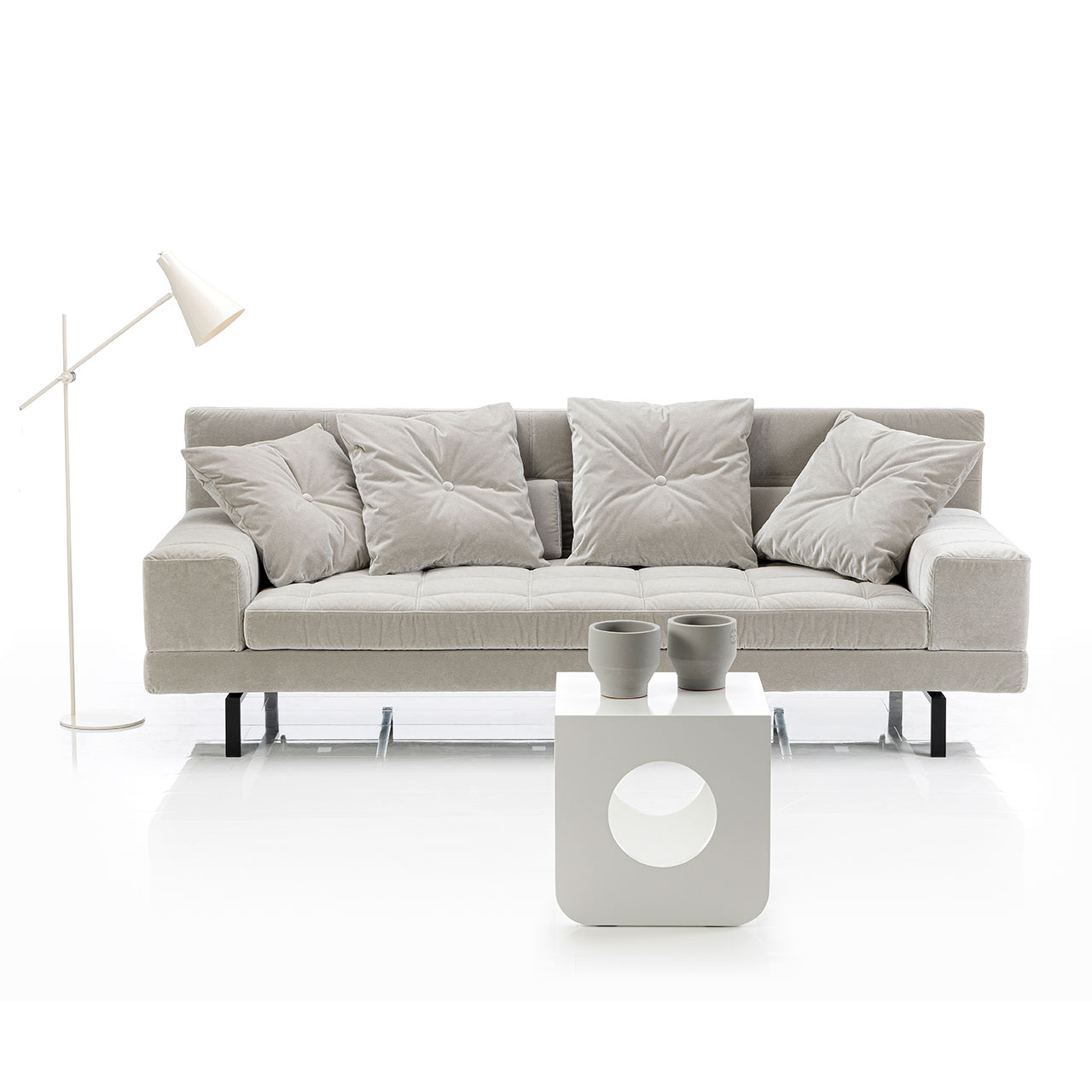 Ambientebild: Graues Sofa mit niedrigen, breiten Armlehnen. Auf dem Sofa liegen vier quadratische Kissen. Vor dem Sofa steht ein weißer Beistelltisch.