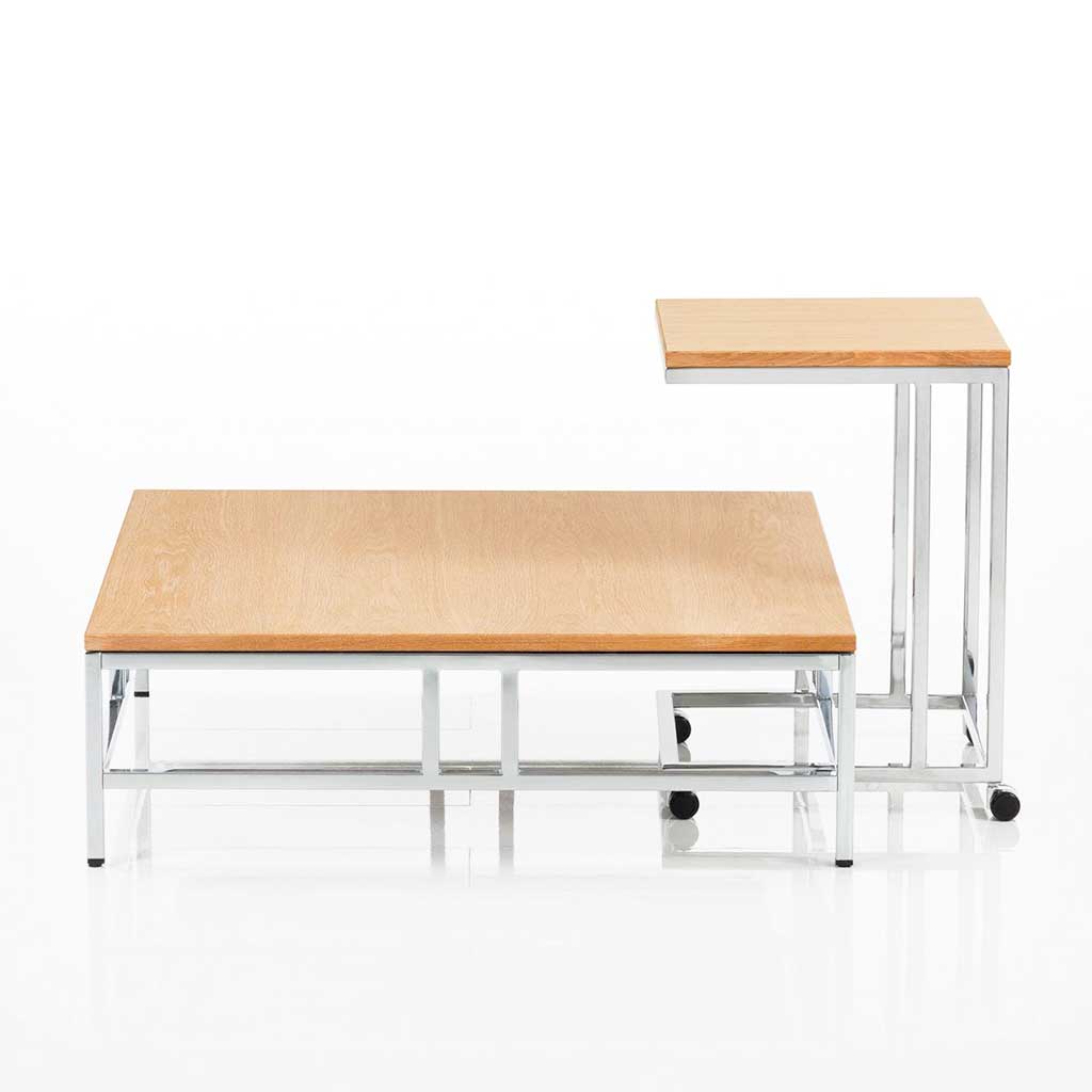 Quadratischer Wohnzimmertisch mit Metall glänzendem Untergestell und Tischplatte in Buche. An den Wohnzimmertisch ist ein höherer Beistelltisch mit Rollen angestellt.