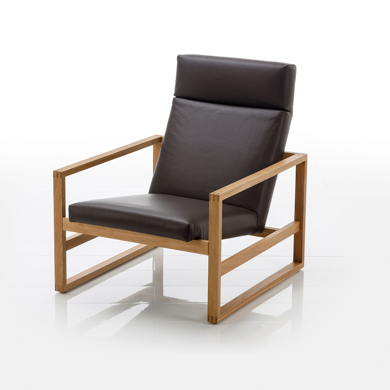 Leicht Schräge Sicht auf einen niedrigen Lounge-Sessel mit Holzgestell in heller Eiche. Die Sitzfläche ist mit schwarzem Leder bezogen.