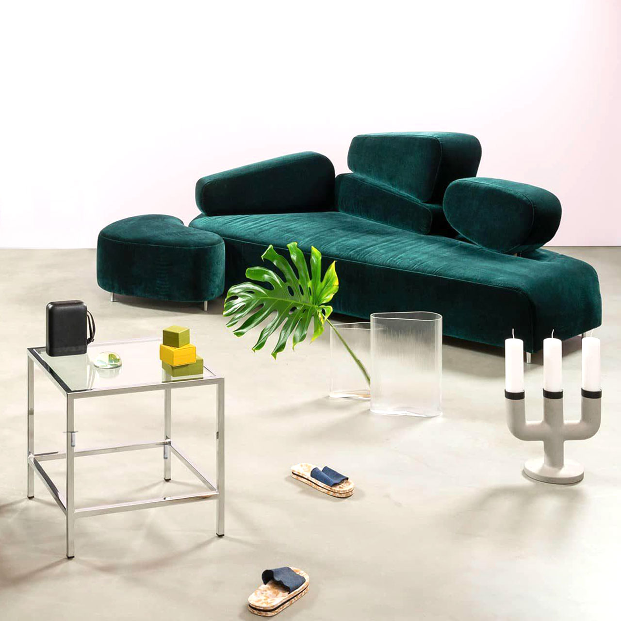 Ambientebild. Grünes asymmetrisches Sofa der Serie brühl mosspink. Links an das Sofa ist ein halbmondförmiger Hocker angestellt. Die Rückenlehne des Sofas besteht aus vier asymmetrischen Elementen.