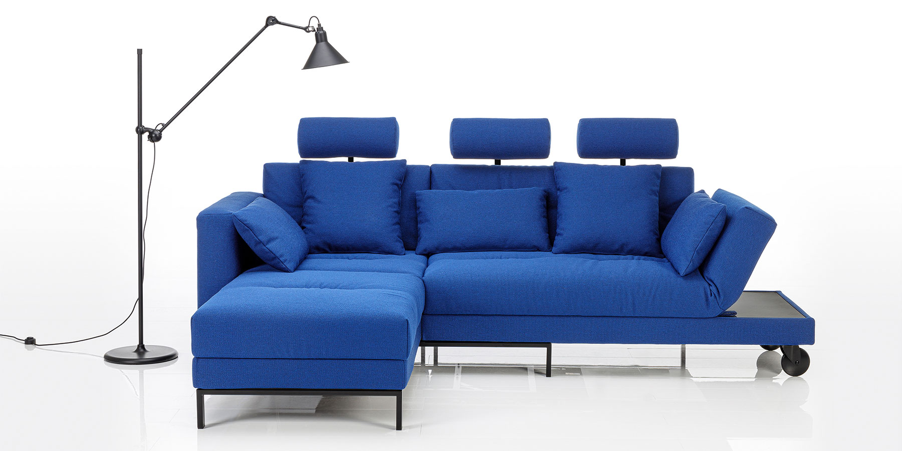 Eine blaue Eckgarnitur aus einem Longchair und einem Anstellsofa. An allen Sitzplätzen ist eine Kopfstütze montiert.