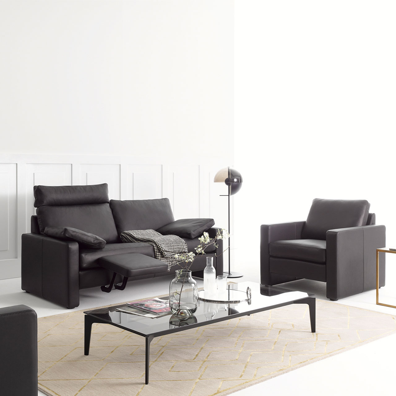 Schwarzes Ledersofa mit breiter Kopfstütze und hochgeklappter Fußstütze. Rechts daneben steht ein Sessel im gleichen schwarzen Leder.