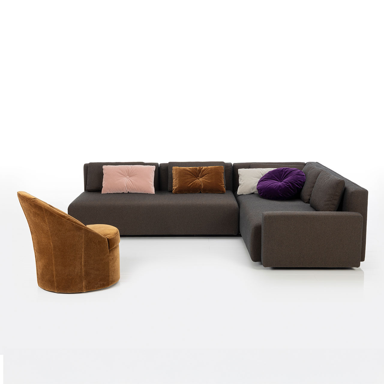 Bodennahes braunes Eckofa mit Drehsofa links und Longchair rechts. Auf dem Sofa liegen drei eckige und ein rundes Kissen. Im Vordergrund steht ein karamellfarbener Sessel.