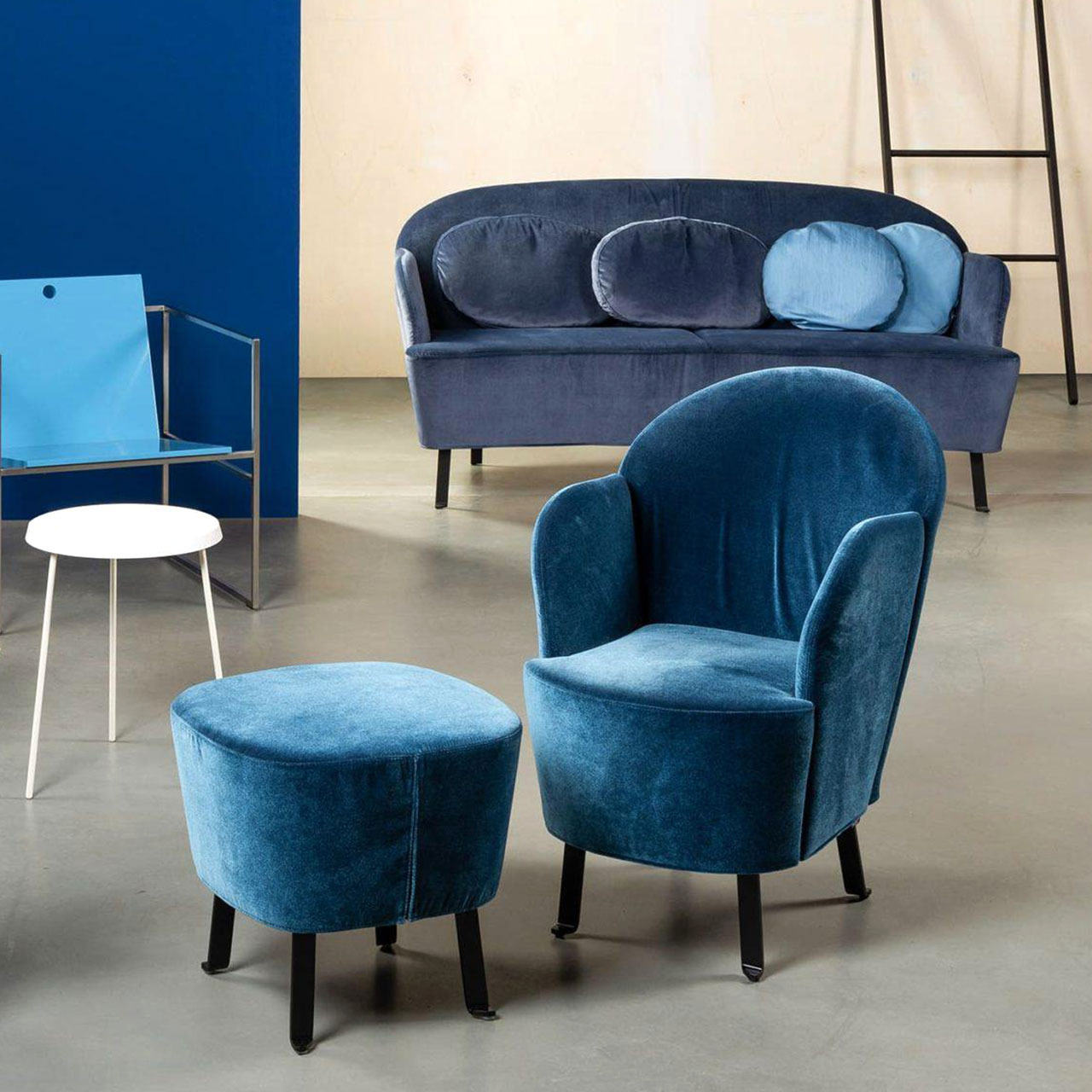 Blauer Sessel mit Füßen in schwarzem Flachstahl mit dazu passendem Hocker. Im Hintergrund steht das dazu passende Sofa in einem etwas dunkleren blauen Stoff.