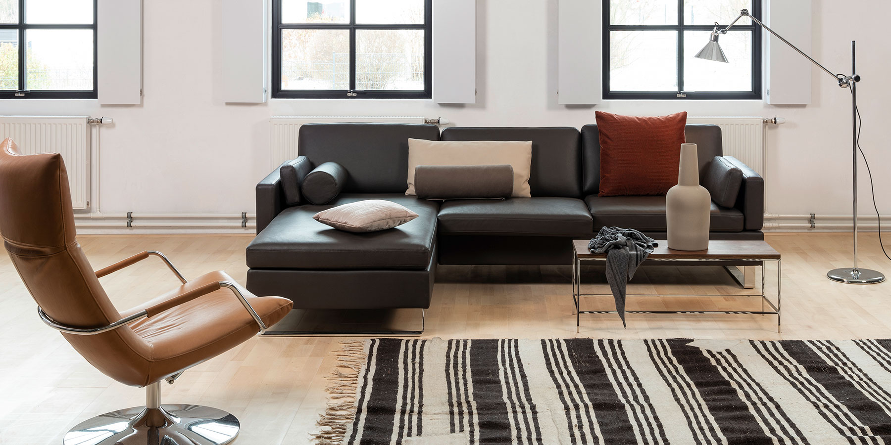 Eine Eckcouch in schwarzem Leder. Auf der Couch liegen Kissen und Kissenrollen in verschiedenen Größen und in Stoff und Leder drapiert.