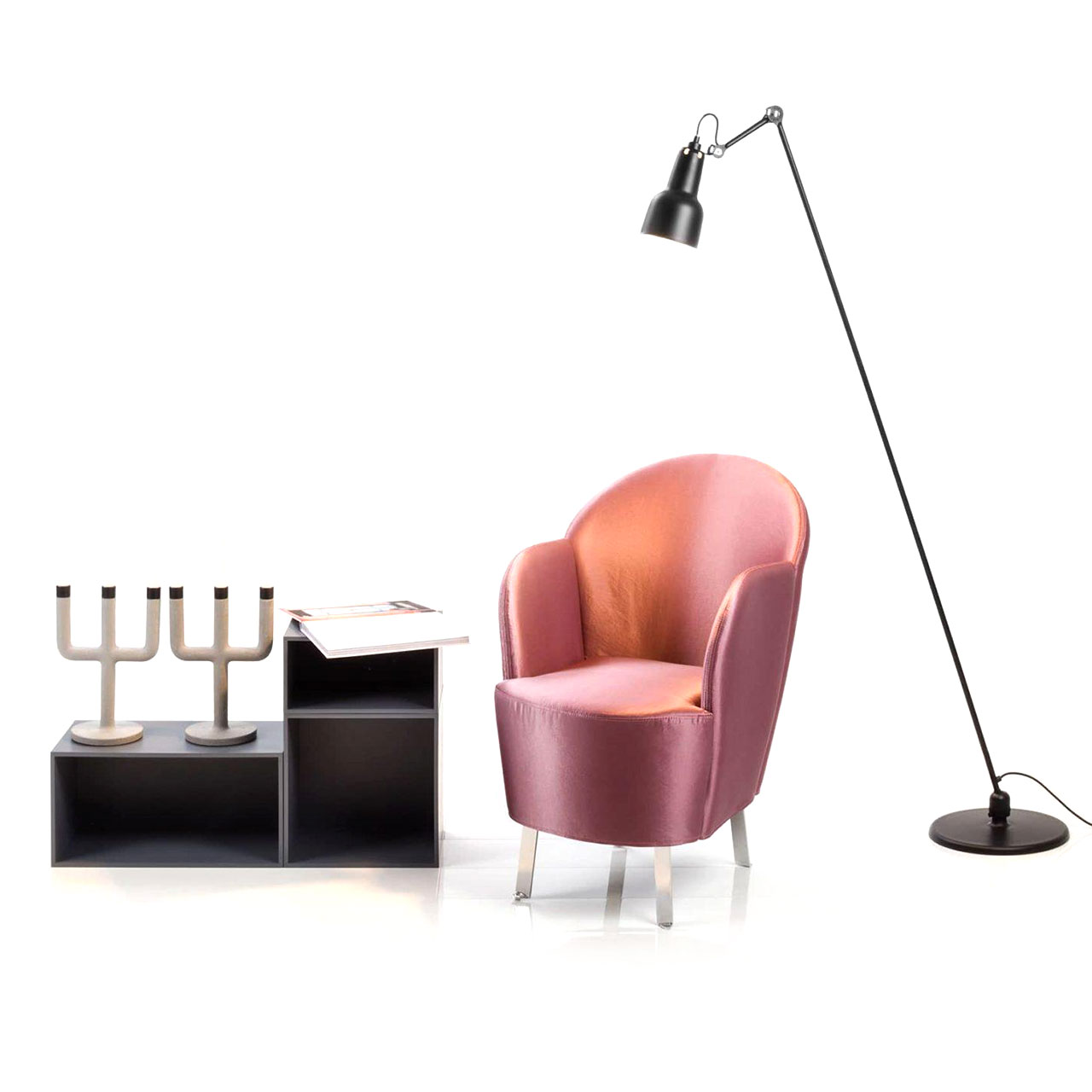 Ein Sessel mit Füßen aus verchromten Flachstahl und mit einem glänzenden Stoff in der Farbe Rose bezogen steht zwischen einem niedrigen Regal und einer Stehlampe.