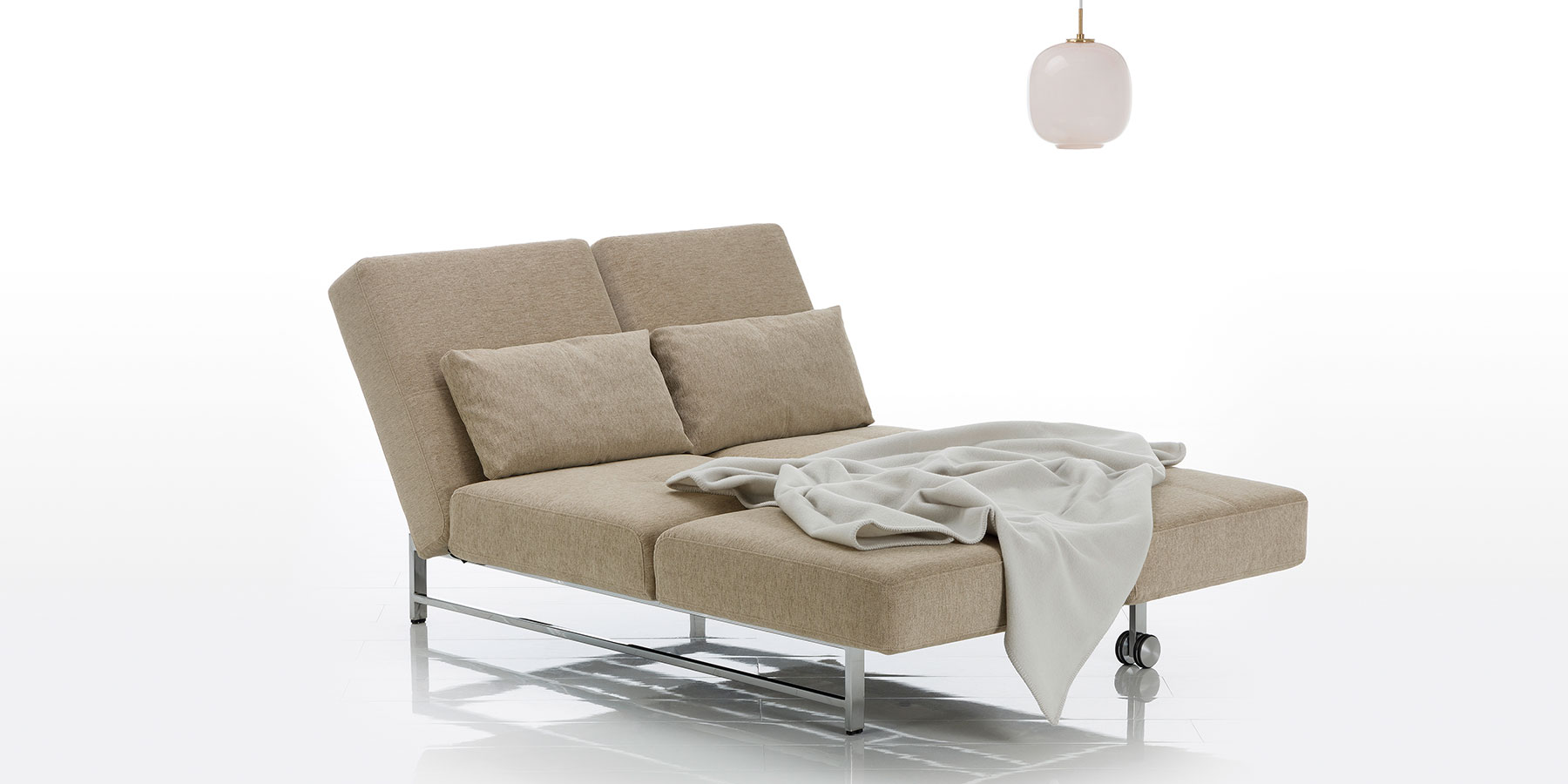 Ein sandfarbenes Sofa, bei dem der Rücken zur Liegefläche abgeklappt wurde. Die links Seite ist hochgeklappt, sodass ein doppeltes Daybed entstanden ist. Auf dem Daybed liegt eine weiße Decke.