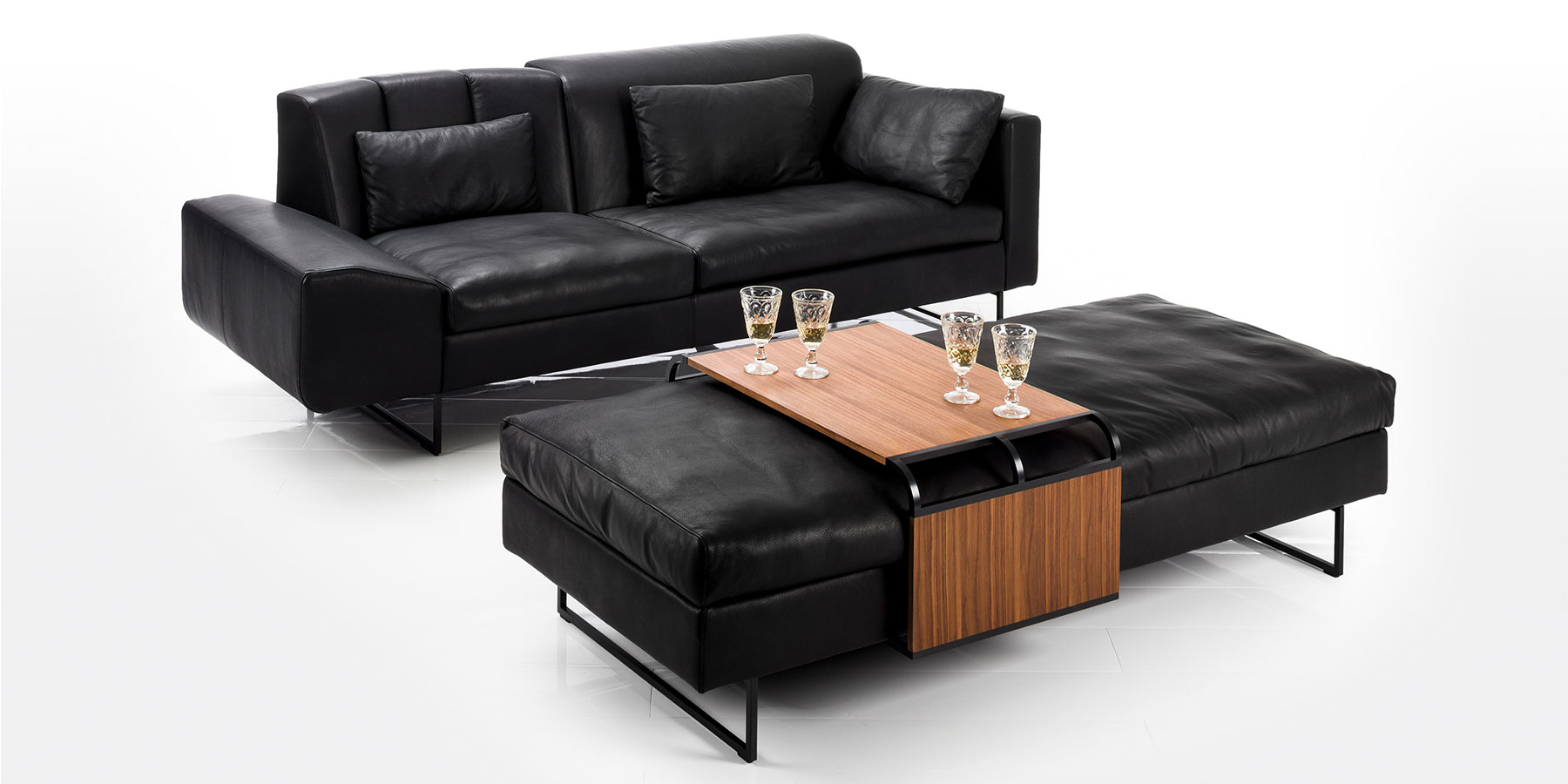 Eine schwarze Ledercouch und eine breite Sitzbank in schwarzem Leder. Auf der Bank liegt ein verschiebbarer Tisch aus dunklem Holz.