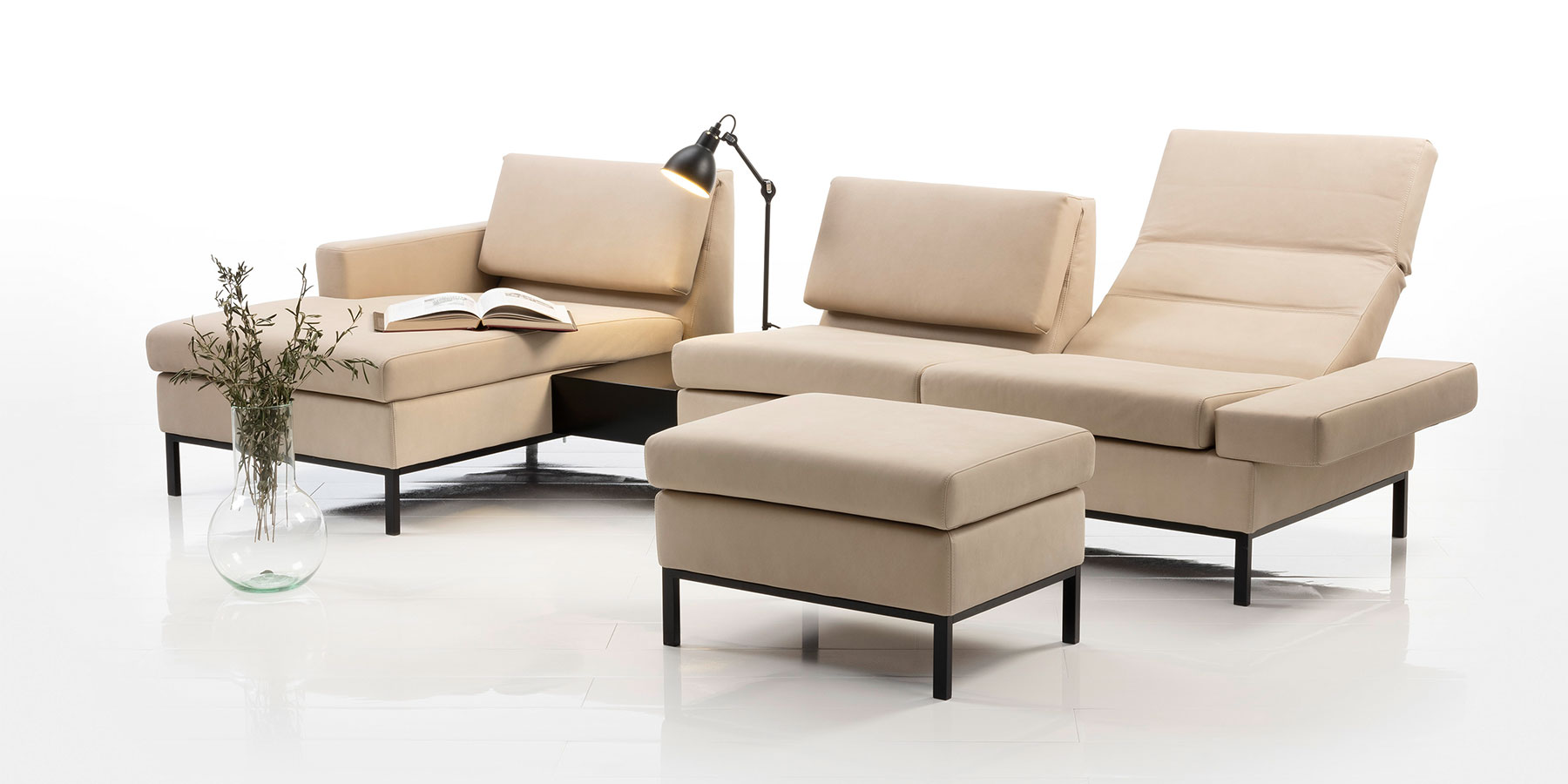 Sandfarbene Eckgarnitur aus Longchair, Ablage und Anstellsofa mit Füßen in dunklem Holz. Im Vordergrund steht ein dazu passender Hocker.