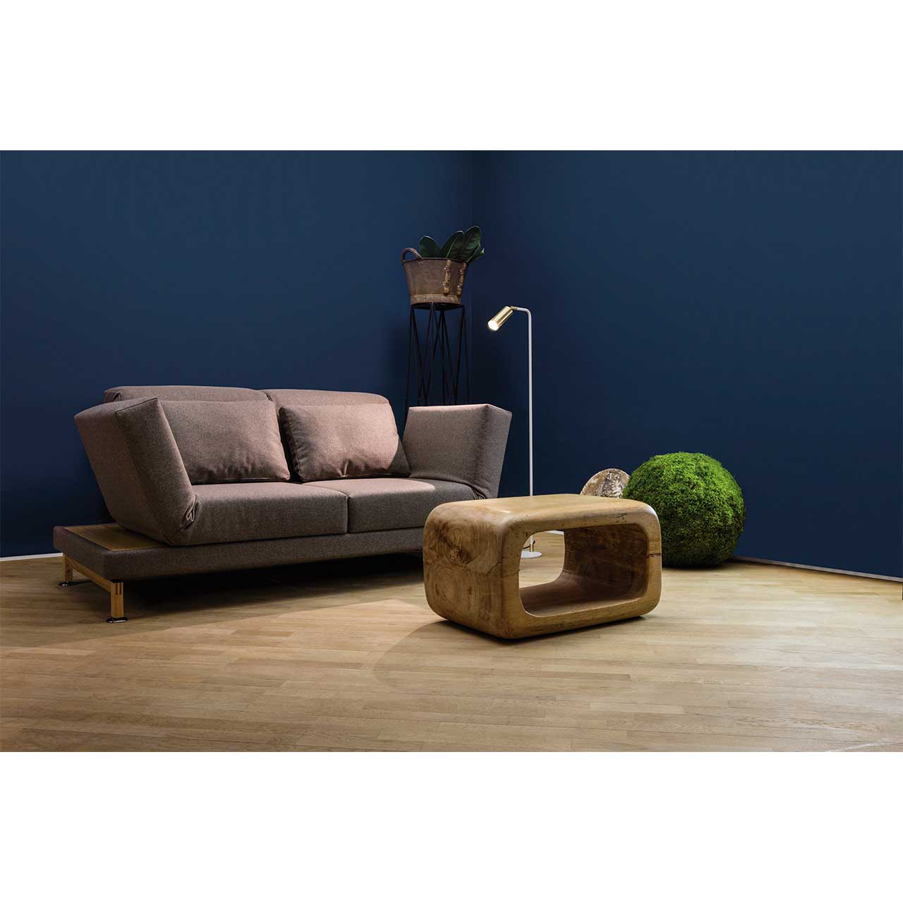Ambientebild. Braunes Stoffsofa mit Kufen in Holz. Rechts und links neben den Armlehnen befinden sich schmale Ablageflächen in hellem Holz. Rechts neben dem Sofa steht eine Stehlampe, vor dem Sofa steht ein Couchtisch aus Holz.
