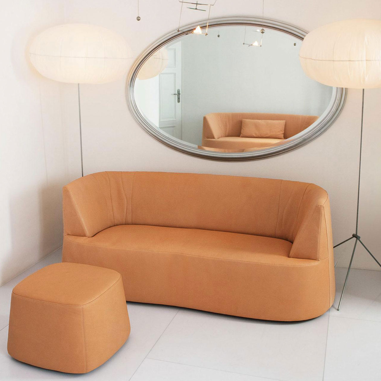 Ambientebild: Ockerfarbenes Sofa mit passendem Hocker vor einer weißen Wand. An der Wand hängt ein ovaler Spiegel mit silbernen Rahmen. 