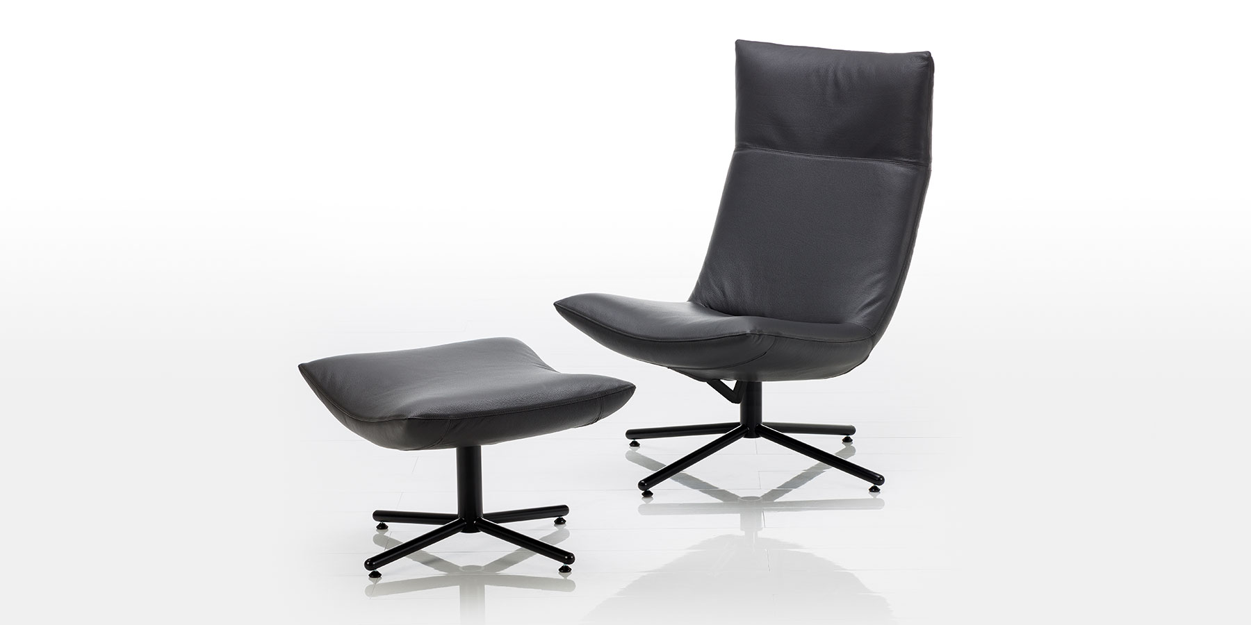 Ein Drehsessel aus schwarzem Leder ohne Armlehnen mit einem schwarzen Kreuzfuß. Daneben steht der passende Hocker, ebenfalls in schwarzem Leder und auf einem schwarzen Kreuzfuß. Die Sitzflächen sind nach unten gewölbt.