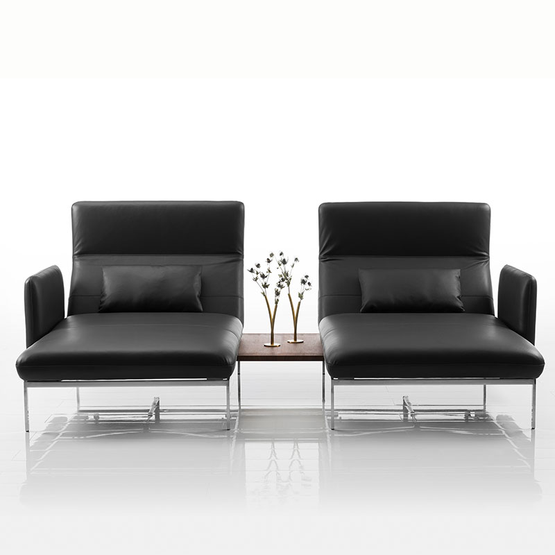 Zwei lederbezogene Longchairs in schwarz mit grazilem Untergestell in Chrom glänzend. Die Longchairs sind durch einen Einhängetisch in dunklem Holz verbunden. Auf dem Tisch stehen zwei Vasen mit filigranen Blumenarrangements.