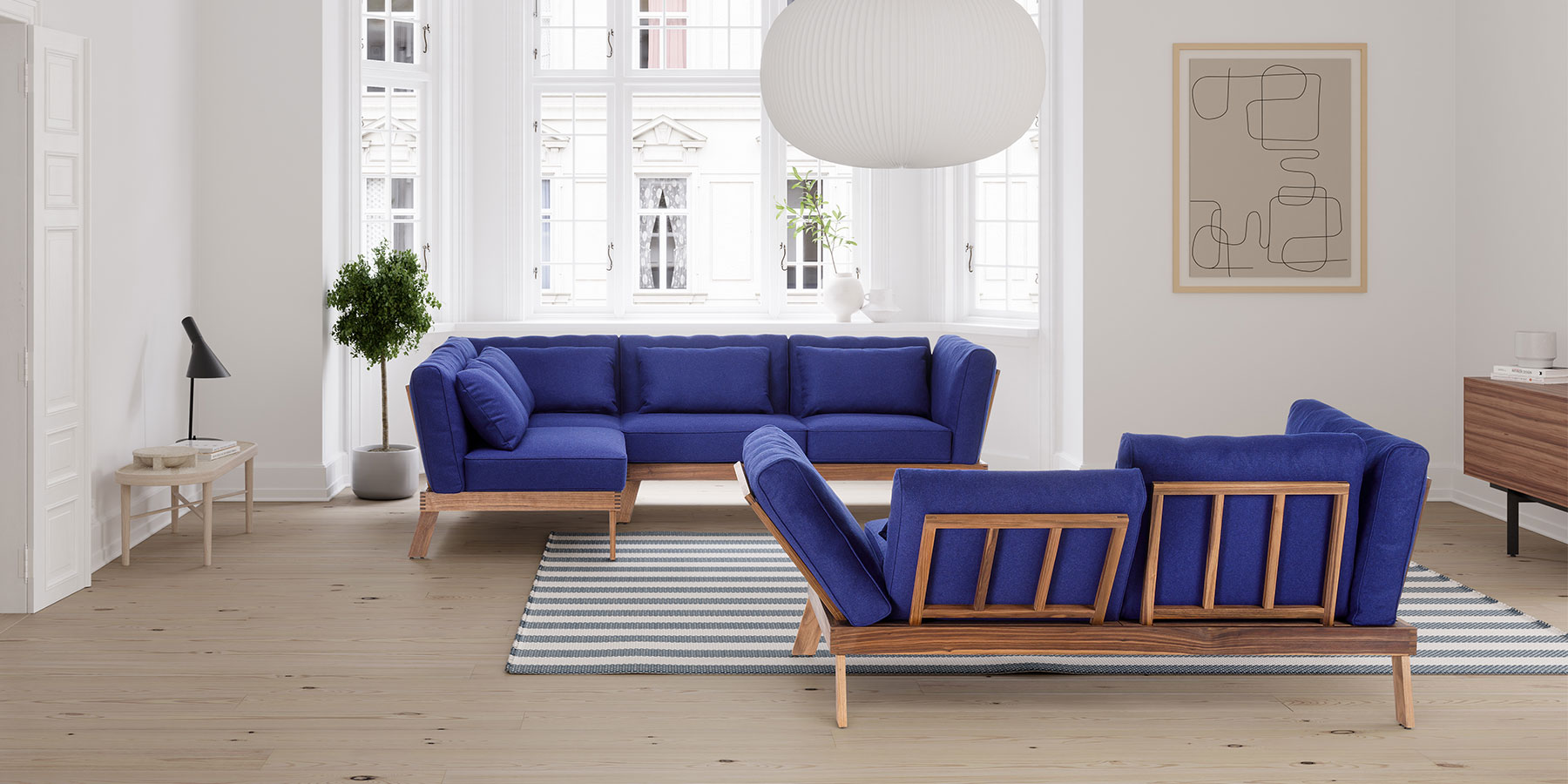 Eine Eckarnitur und ein Sofa stehen einander leicht schräg versetzt gegenüber. Die aufgelegten Polster sind dunkelblau, das Holzgestell aus heller Eiche.