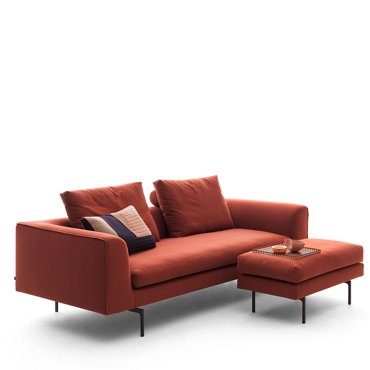 Rotes Sofa auf schwarzen Einzelfüßen mit drei Kissen dekoriert, davor ein Hocker im passenden Design.