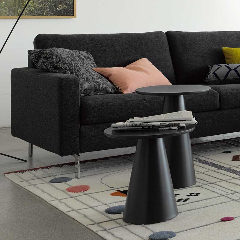 schwarze Beistelltische in zwei Höhen mit konischem Fuß und runder Platte vor schwarzem Sofa