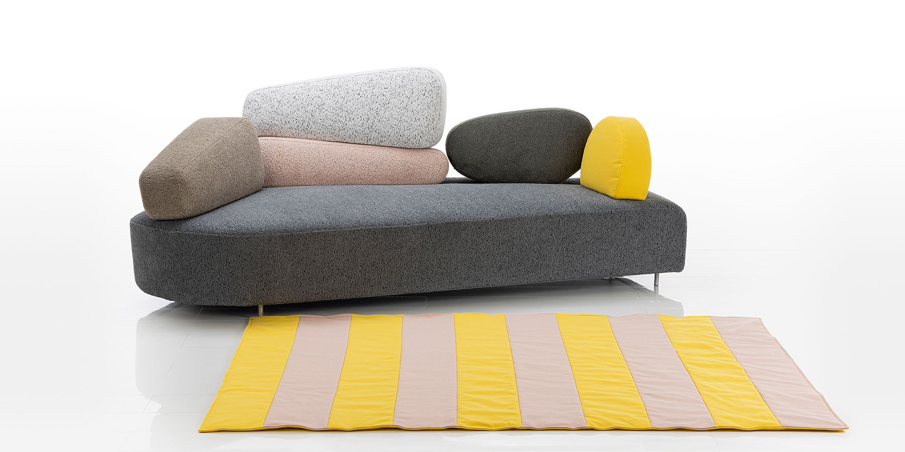 Ein asymmetrisch geformtes Sofa mit einer Sitzbasis aus grauem Stoff. Rücken und Seiten bestehen aus sechs unregelmäßig geformten Segmenten in den Farben braun, rosé, hellgrau, dunkelgrau und gelb.