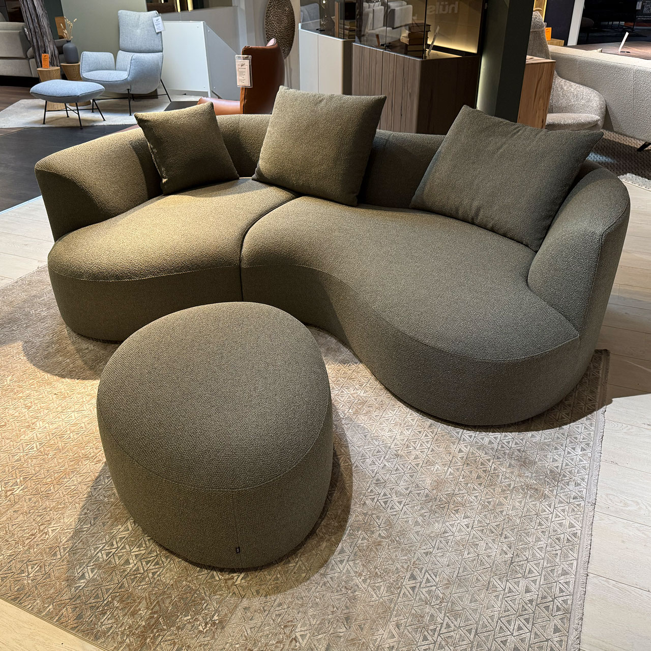 Organisch geformtes Sofa aus graugrünem Stoff mit Liege- und Sitzteil mit einem kleinen und zwei großen Kissen, davor ein ovaler Hocker