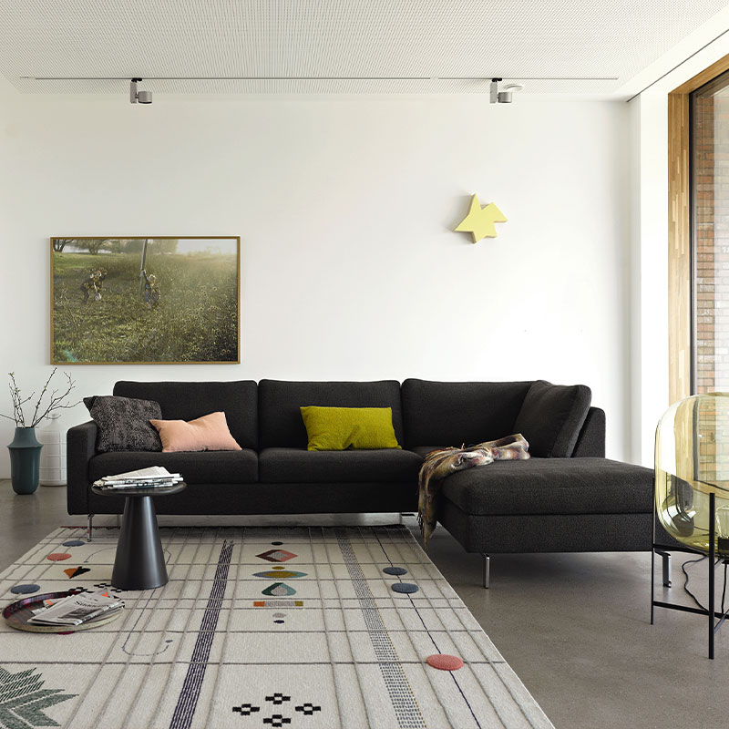 Stoffbezogene Eckcouch vor einer weißen Wand. Der Stoff ist grün-braun, auf der Garnitur liegen Kissen.