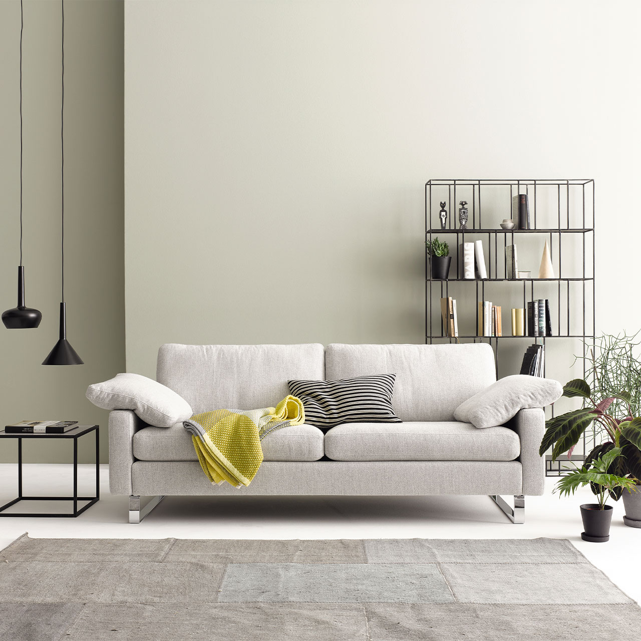 Ambientebild des Sofas Conseta von COR und beigem Stoff. Auf dem Sofa sind drei Kissen und eine Decke drapiert.