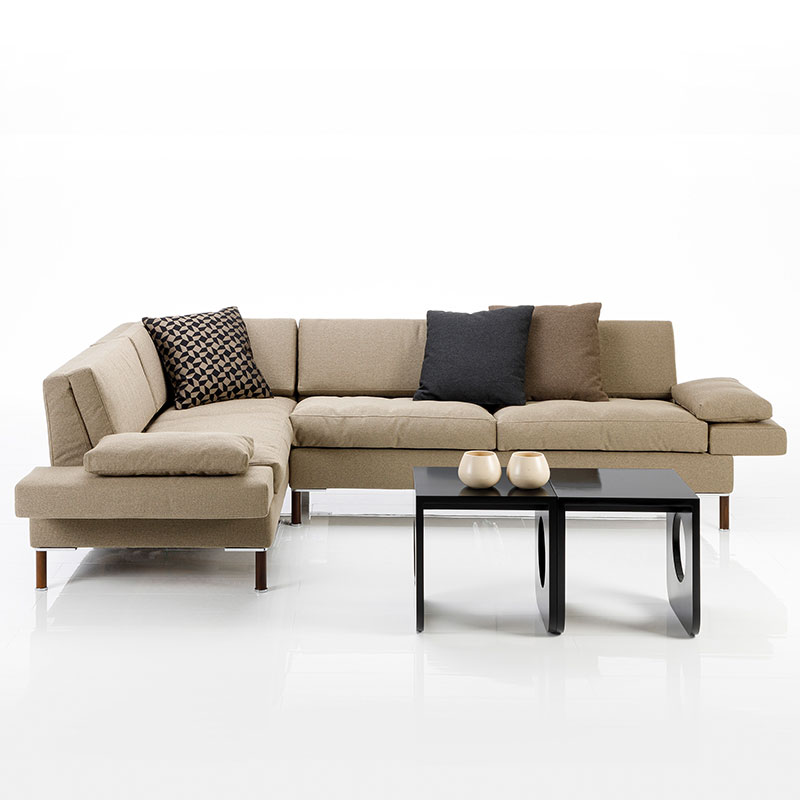 Eckgarnitur in hellbraunem Stoff mit gemusterten Kissen. Vor der Kombination stehen zwei schwarzen Beistelltische.