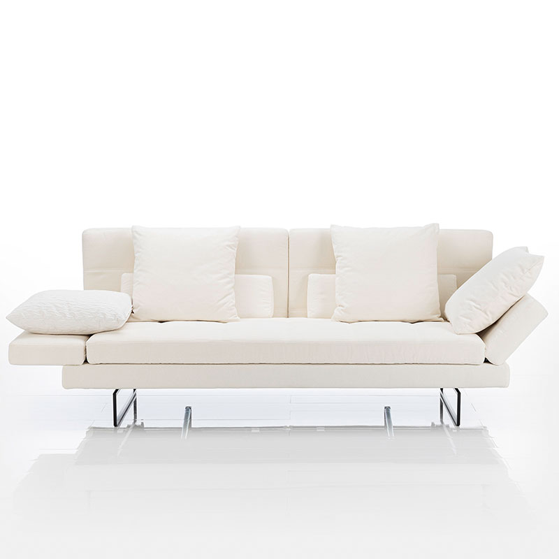 Ein weißes Sofa mit Untergestell in Edelstahl. Die linke Armlehne ist ganz abgesenkt, die rechte halb. Auf dem Sofa liegen 6 Kissen.