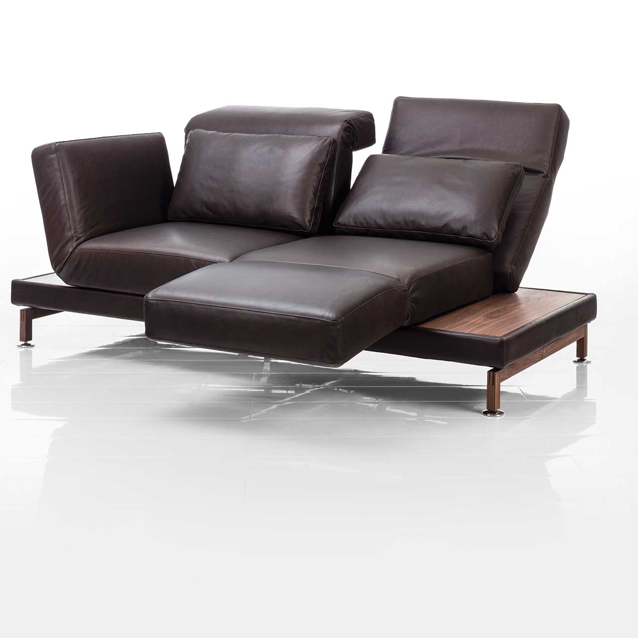 Dunkelbraunes Ledersofa. Rechts und links neben den Armlehnen befinden sich schmale Ablageflächen in dunklem Holz. Die rechte Armlehne ist als Fußsütze vorgeschwenkt, die Rückenlehne leicht abgesenkt.
