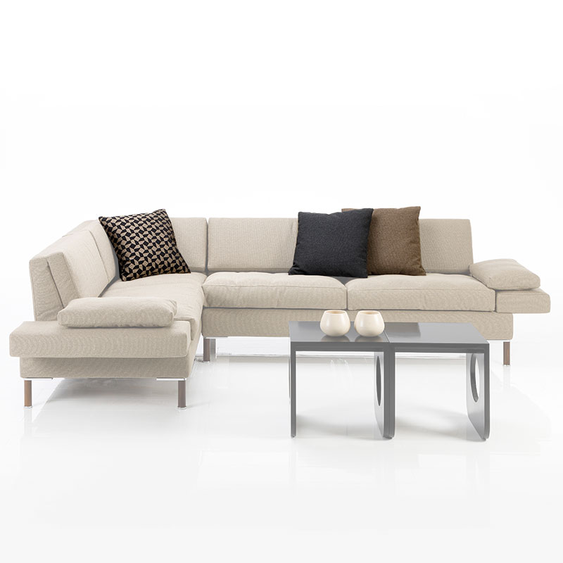 Eckgarnitur in hellbraunem Stoff mit gemusterten Kissen. Vor der Kombination stehen zwei schwarzen Beistelltische. Die Kissen sind farblich hervorgehoben.