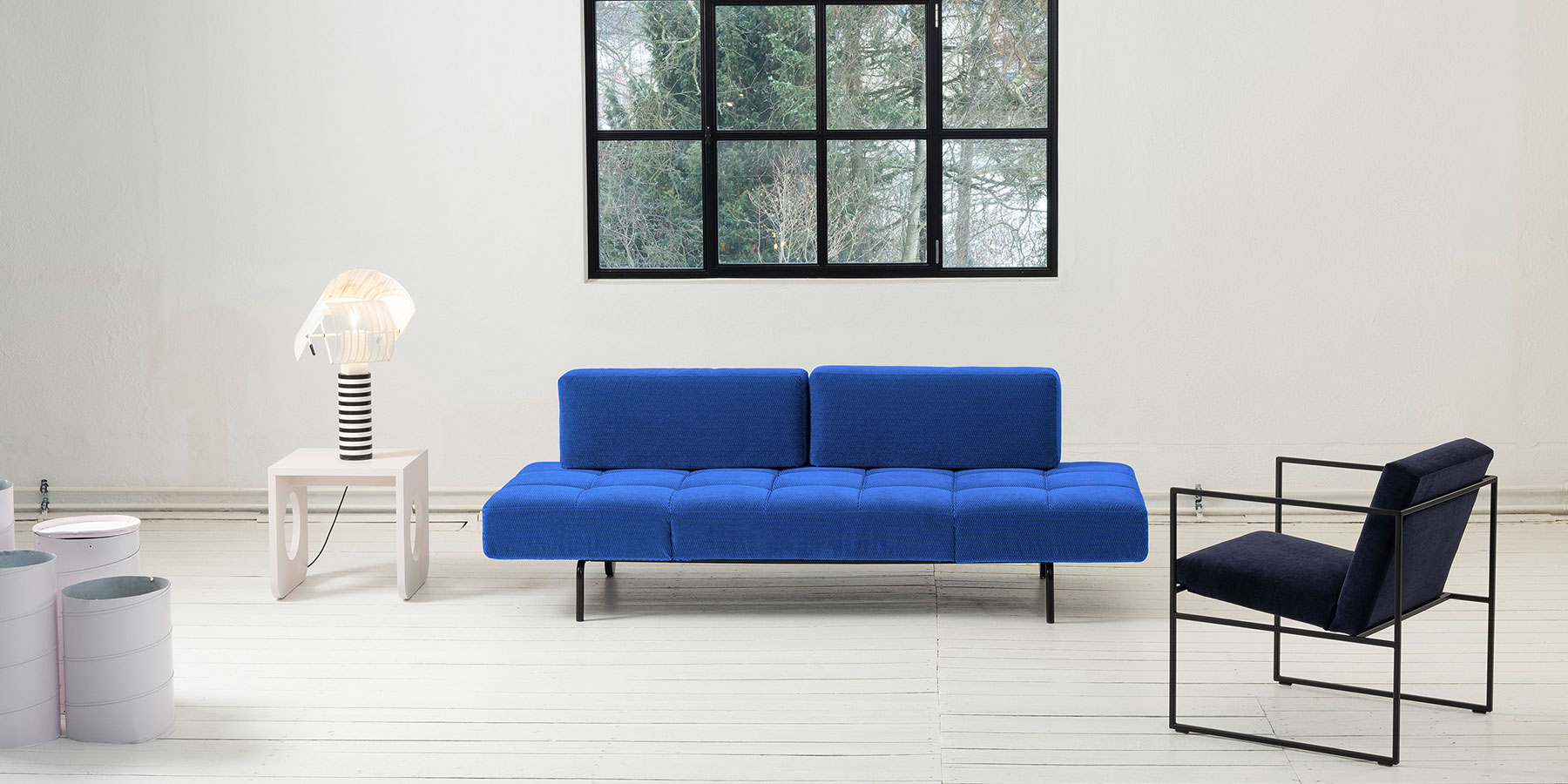 Ein blaues Sofa ohne Armlehnen. Die Sitzfläche ist quadratisch gesteppt. Die zwei länglichen Lehnenpolster sind glatt gearbeitet und kürzer als die Sitzfläche. Das Metallgestell ist schwarz. 