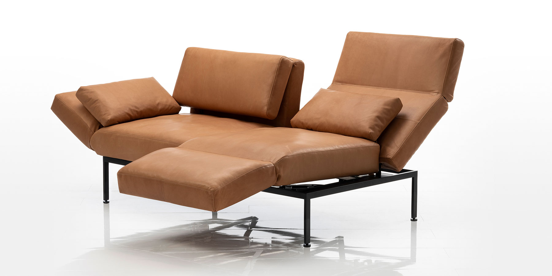 Ein zweisitziges Sofa in braunem Leder. Am rechten Sitzplatz wurde die Armlehne zur Fußstütze umgeschwenkt, um eine Relaxposition herzustellen. Die linke Armlehne wurde leicht abgesenkt.