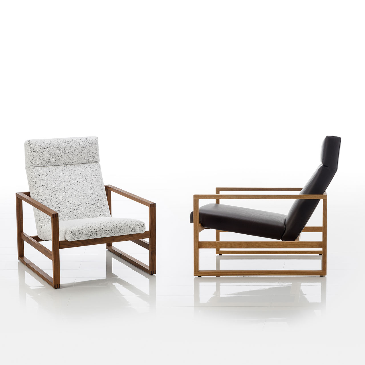 Zwei Lounge-Sesse. mit nieriger Sitzhöhe. Die Gestelle sind aus Holz. Der linke Sessel ist mit einem weißen, strukturierten Stoff bezogen, der rechte Sessel ist mit schwarzem Leder bezogen.