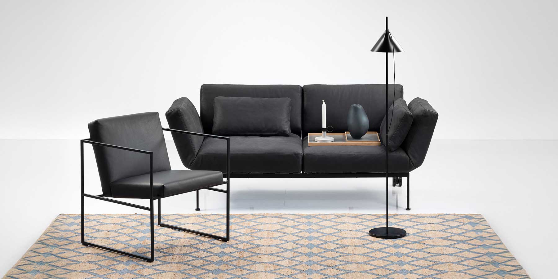Schwarzes Ledersofa mit einem Rückenkissen. Auf der rechten Sitzfläche steht ein Tablett mit einer Kerze und einer Vase. Vor dem Sofa stehen eine schwarze grazile Stehlampe rechts und ein mit schwarzem Leder bezogener Stuhl.