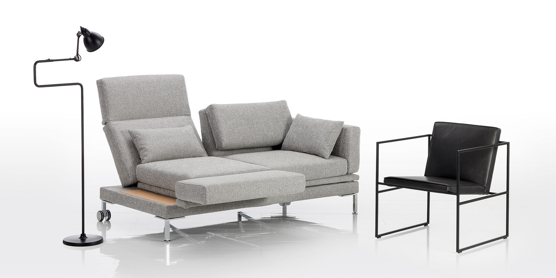 Ein Sofa in grauem Stoff. Die linke Rüclenlehne ist aufgefaltet und leicht abgesenkt. Die linke Armlehne wurde als Fußstütze nach vorne geschwenkt.