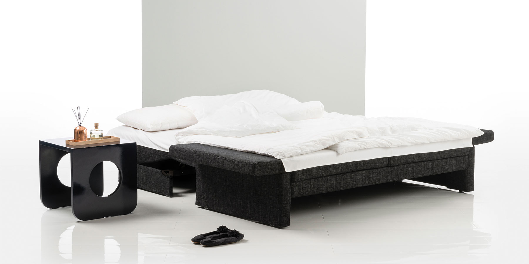 Ein zu einem Doppelbett ausgeklapptes Schlafsofa. Decken und Kissen sind mit weißer Bettwäsche bezogen. Neben dem Bettsofa steht ein Beistelltisch.