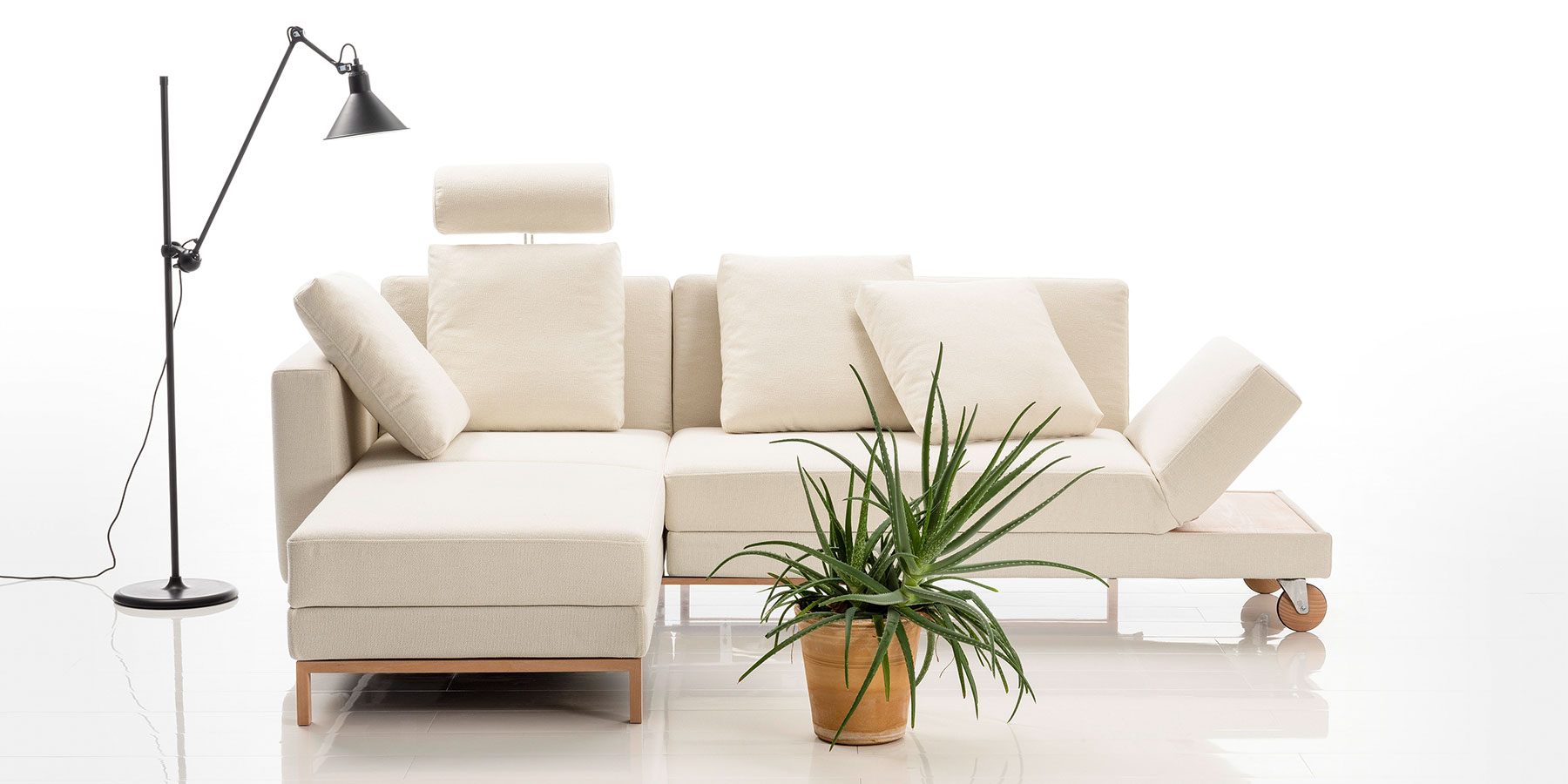 Eine helle Eckgarnitur mit Füßen aus hellem Holz. Auf der linken Seite mit einem schmalen Longchair, rechts mit einem kurzen Anstellsofa.