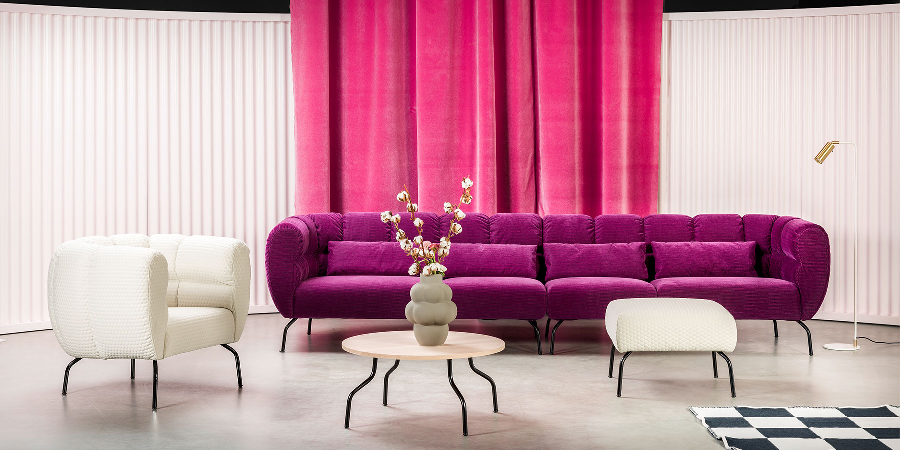 Ein Sessel und ein Sofa stehen im rechten Winkel zueinander. Das Gestell der Polstermöbel sind filigrane, gebogene, schwarze Metallfüße. Die Polster der Möbel sind opulent und umschließen die Sitzbasis ganz.