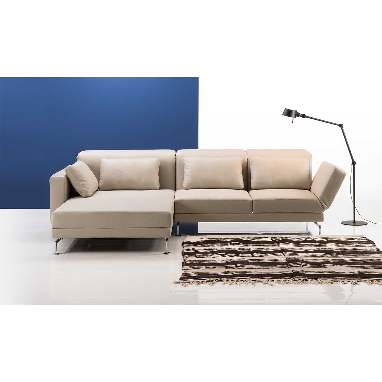 Eckgarnitur mit breitem Longchair links in beigem Leder. Rechts daneben steht eine Lampe, vor der Garnitur liegt ein gestreifter Teppich.