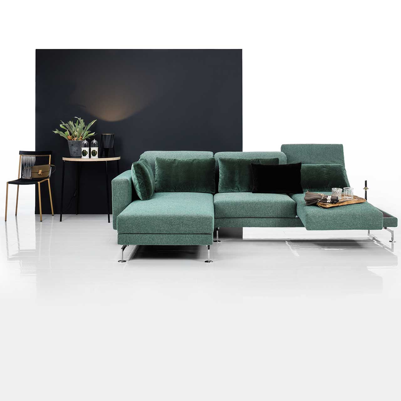 Eckgarnitur mit Longchair links in grünem Stoff. Rechts neben der Armlehne befindet sich eine Ablagefläche in dunklem Holz. Die Armlehne ist zur Relaxposition vorgescchwenkt, die Rückenlehne ist abgesenkt.